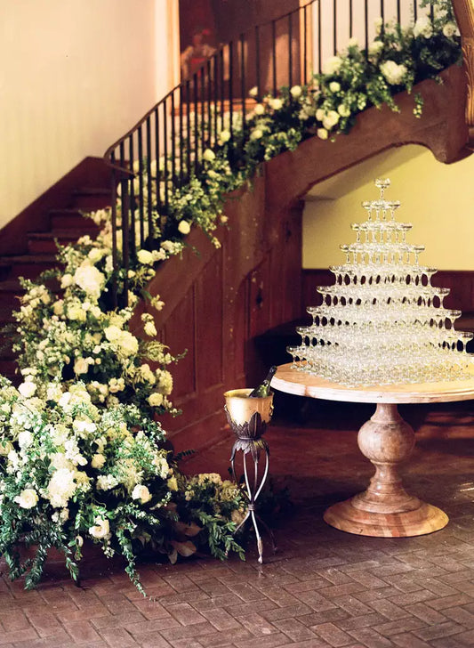 9-tier Champagne Tower set on beautiful round table with floral arrangements behind going up a staircase