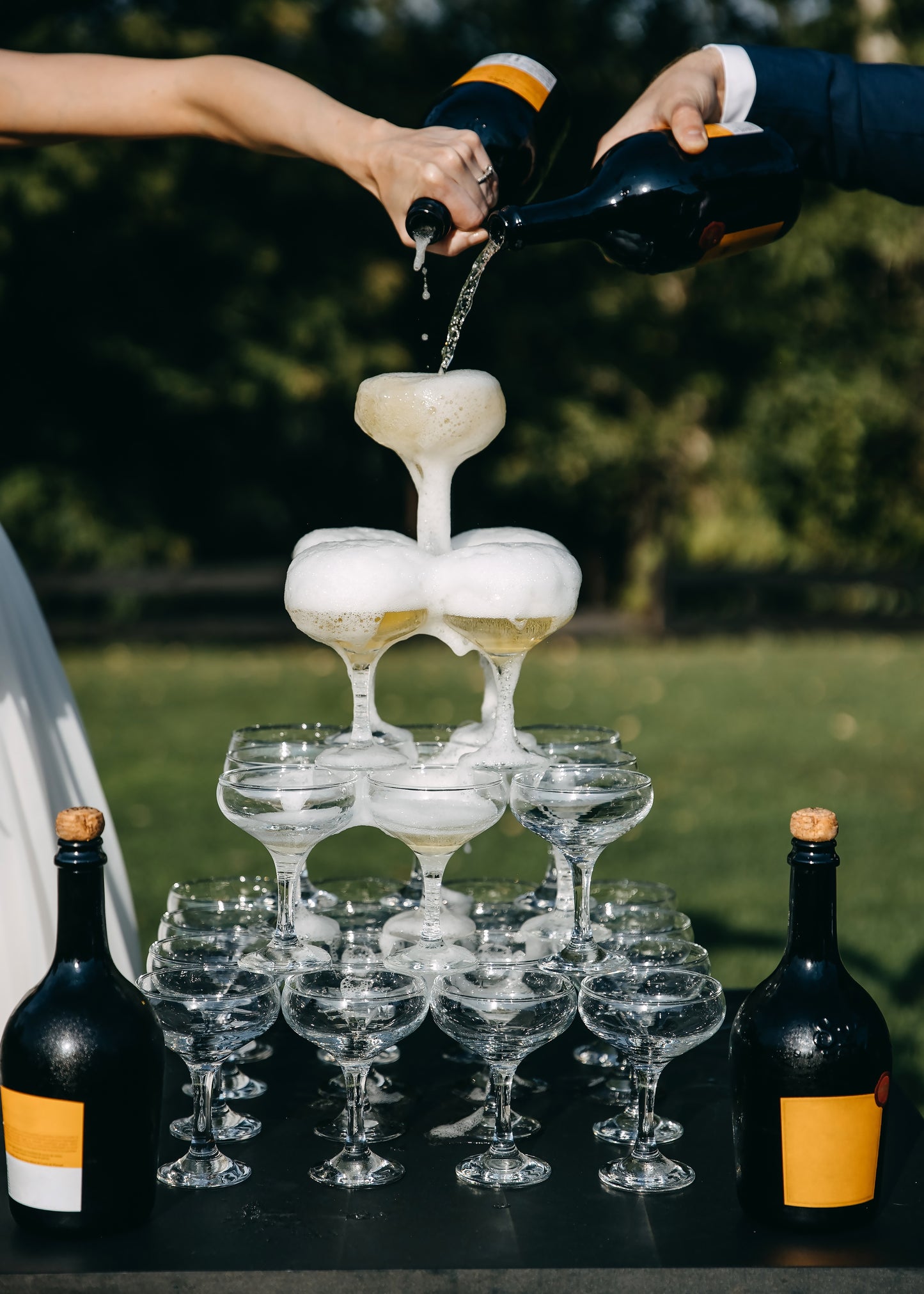 4-tier Champagne Tower with bride and groom both filling the glasses. Set outside in a lush green garden.  